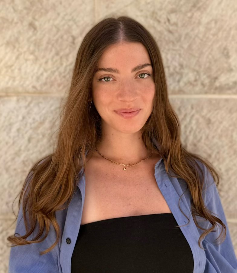 Portrait of a smiling woman with long brown hair, wearing a blue shirt over a black top, standing against a stone wall.