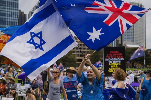 Crowd at an outdoor rally waving Israeli and Australian flags with protest signs in the background.
