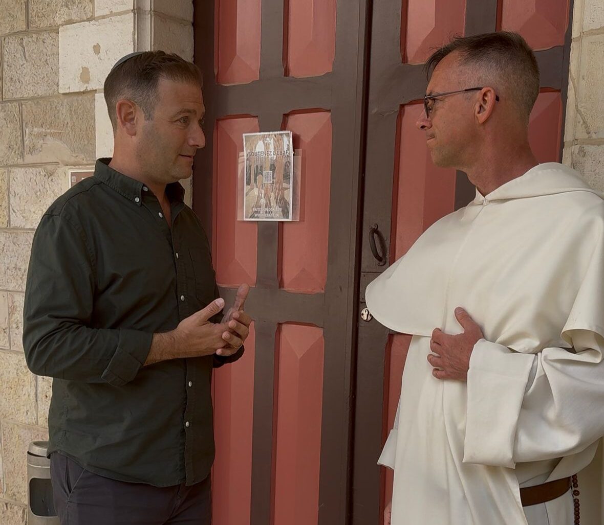 Two men stand facing each other by a large red-and-brown door, one in a dark shirt, the other in a white religious habit, conversing.