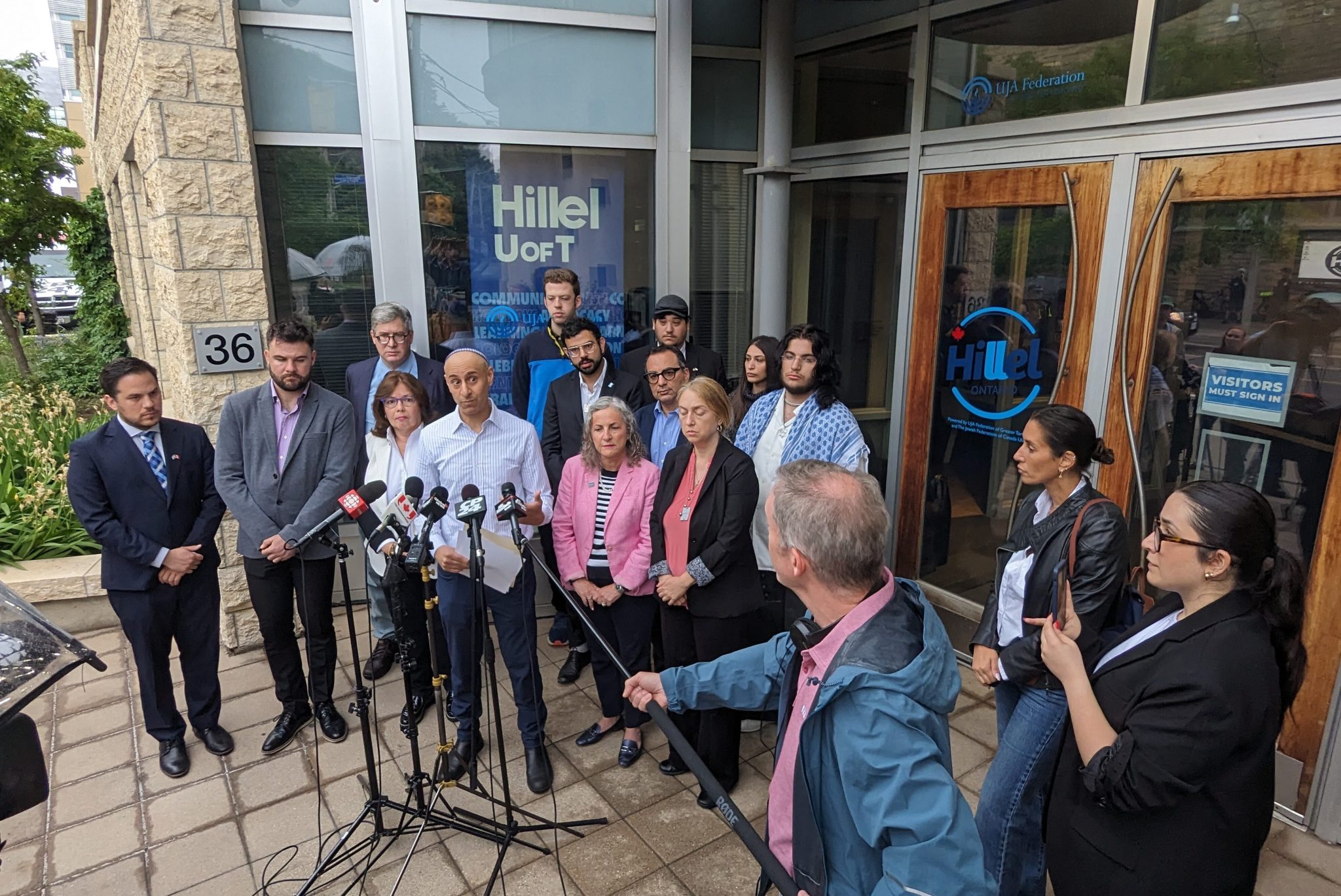 Group of officials and reporters at a outdoor press conference outside a building entrance with multiple microphones on stands gathered in front of them.