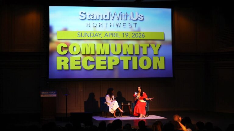Stage scene with two women seated and speaking during a community reception; large screen displays 'StandWithUs Northwest' and 'Community Reception' poster.