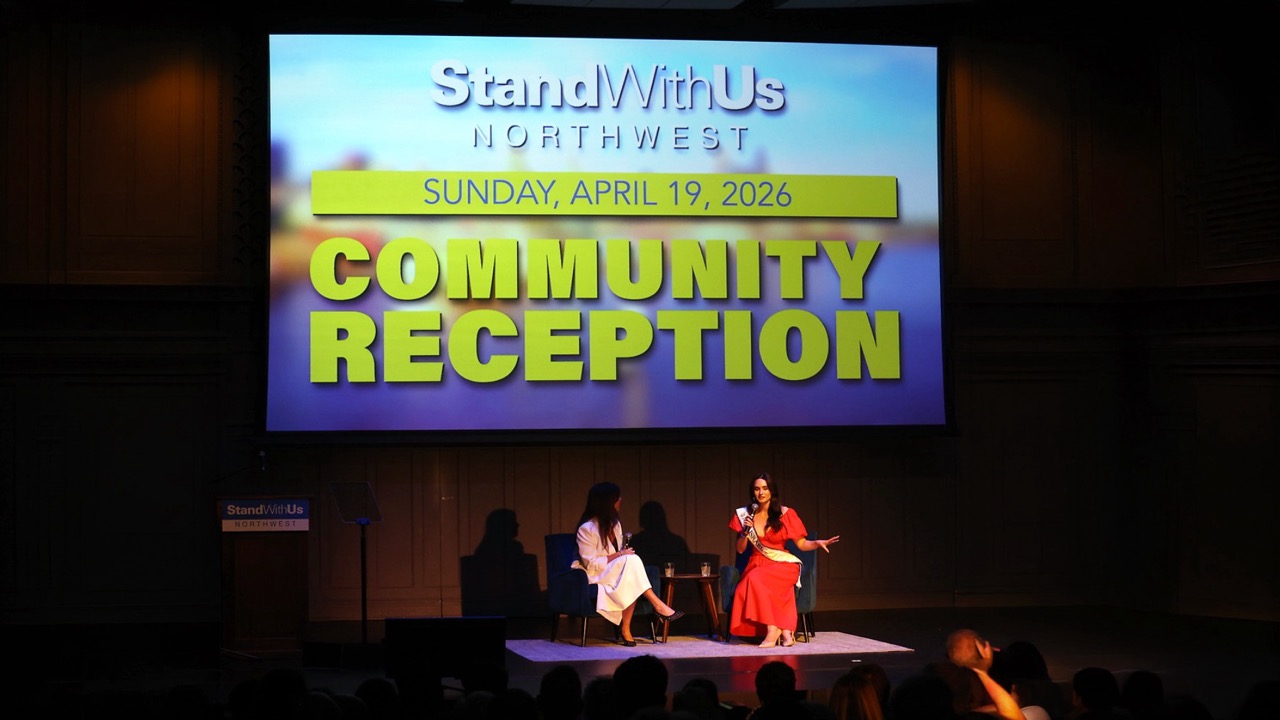 Stage scene with two women seated and speaking during a community reception; large screen displays 'StandWithUs Northwest' and 'Community Reception' poster.