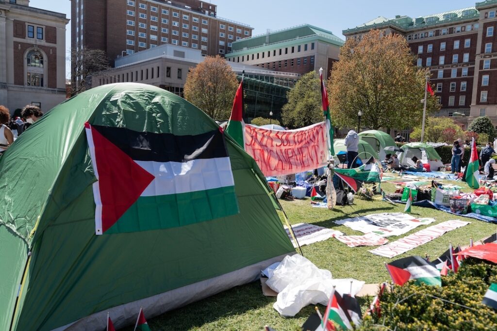 Green camping tent with a Palestinian flag attached, set up on a grassy campus lawn with banners and tents in the background.