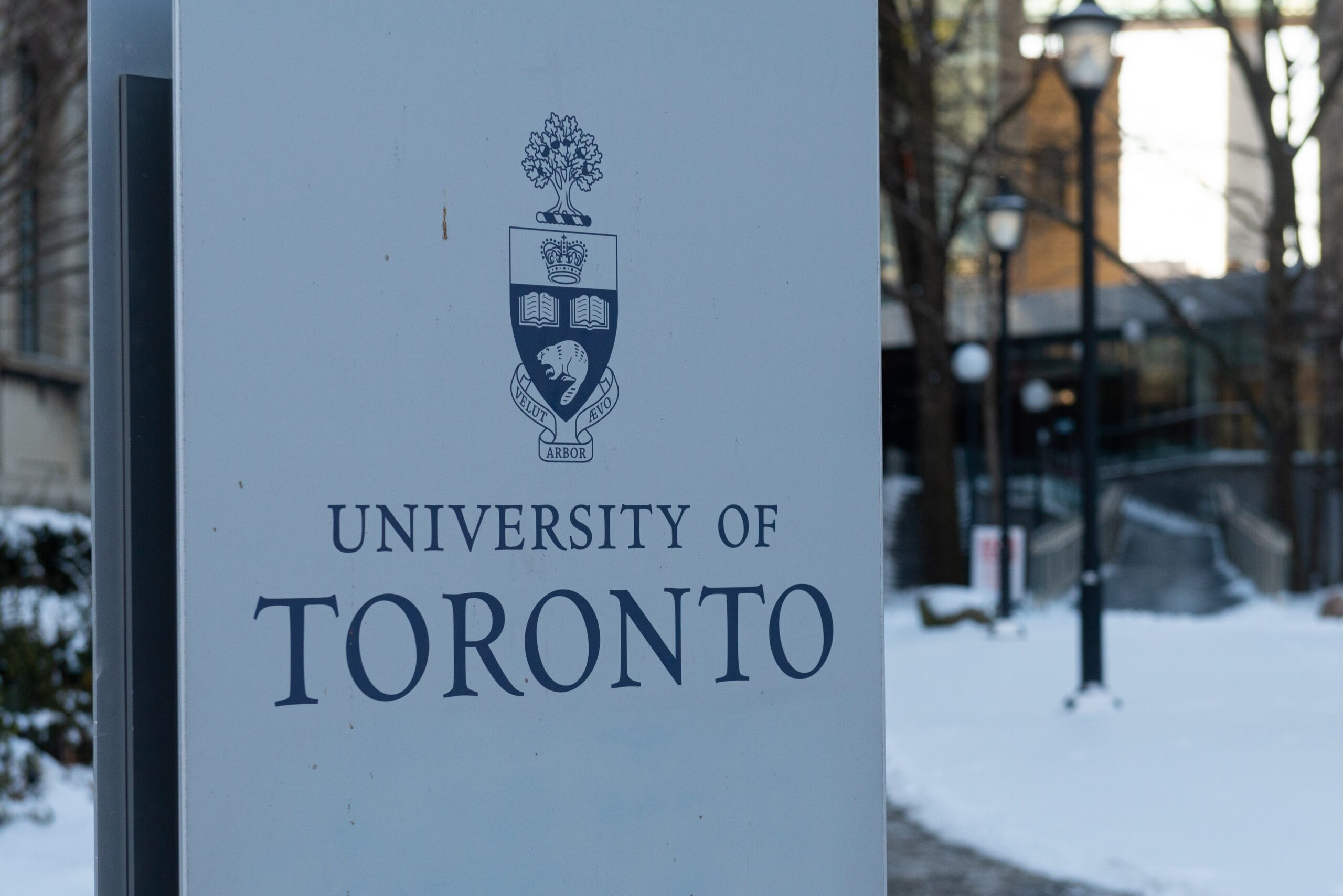 Sign for the University of Toronto with its crest and motto in blue, outdoor snowy scene in the background