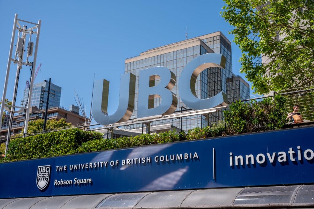 Large metallic UBC letters on a blue sign with a glass-front building and trees in the background at Robson Square, Vancouver.