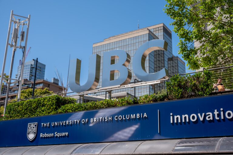 Large metallic UBC letters on a blue sign with a glass-front building and trees in the background at Robson Square, Vancouver.