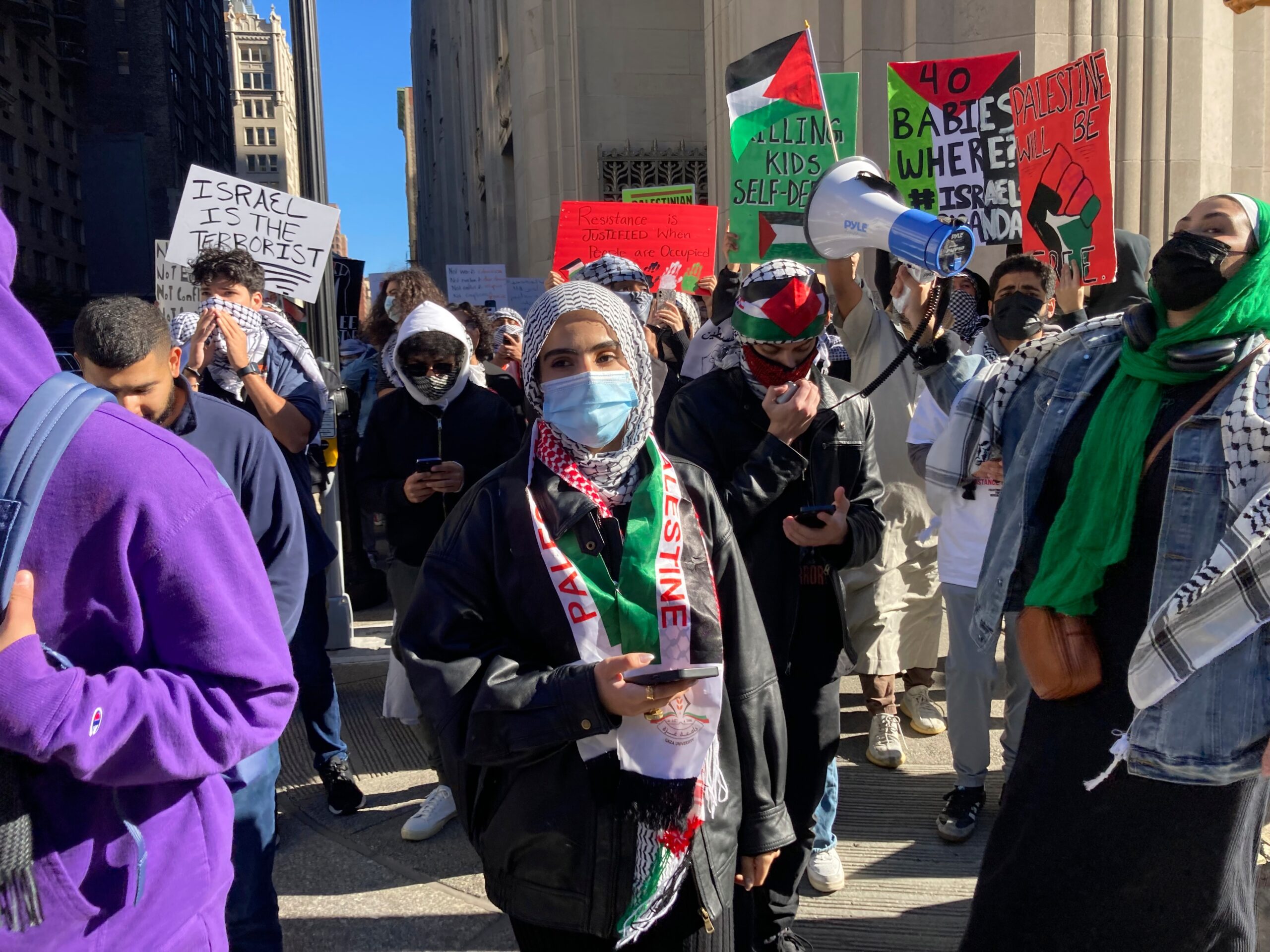 A diverse group of protesters marching on a city street, holding colorful signs and Palestinian flags; many wear keffiyehs and masks.