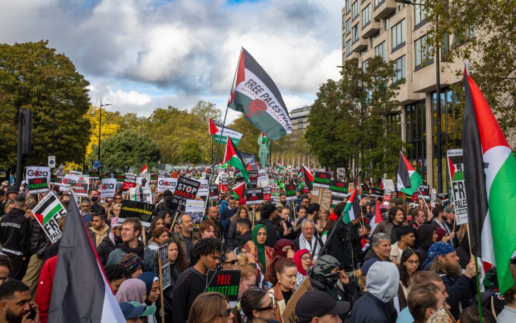 Large street protest with Palestinian flags and signs reading Free Palestine, crowds filling the avenue in front of office buildings.