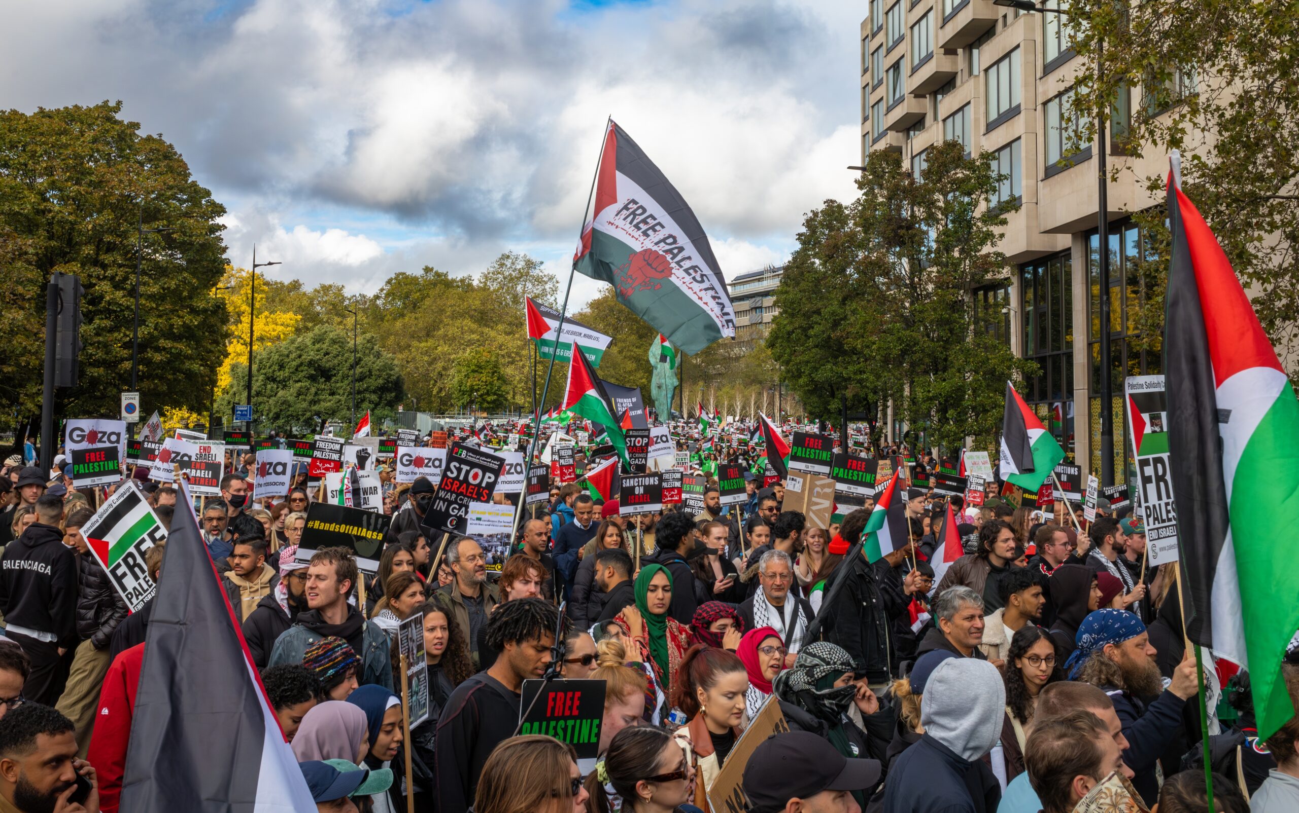 Large street protest with Palestinian flags and signs reading Free Palestine, crowds filling the avenue in front of office buildings.