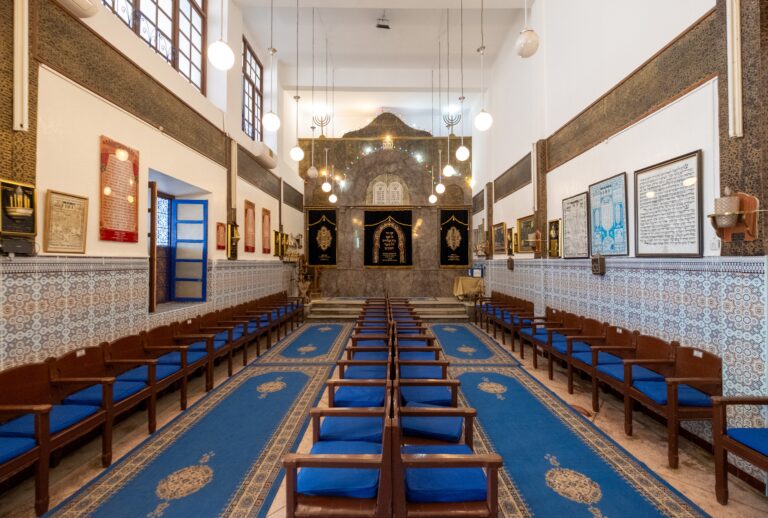 Interior of a synagogue with rows of blue-cushioned wooden benches, a long blue carpet runner, and a decorative ark at the far end.