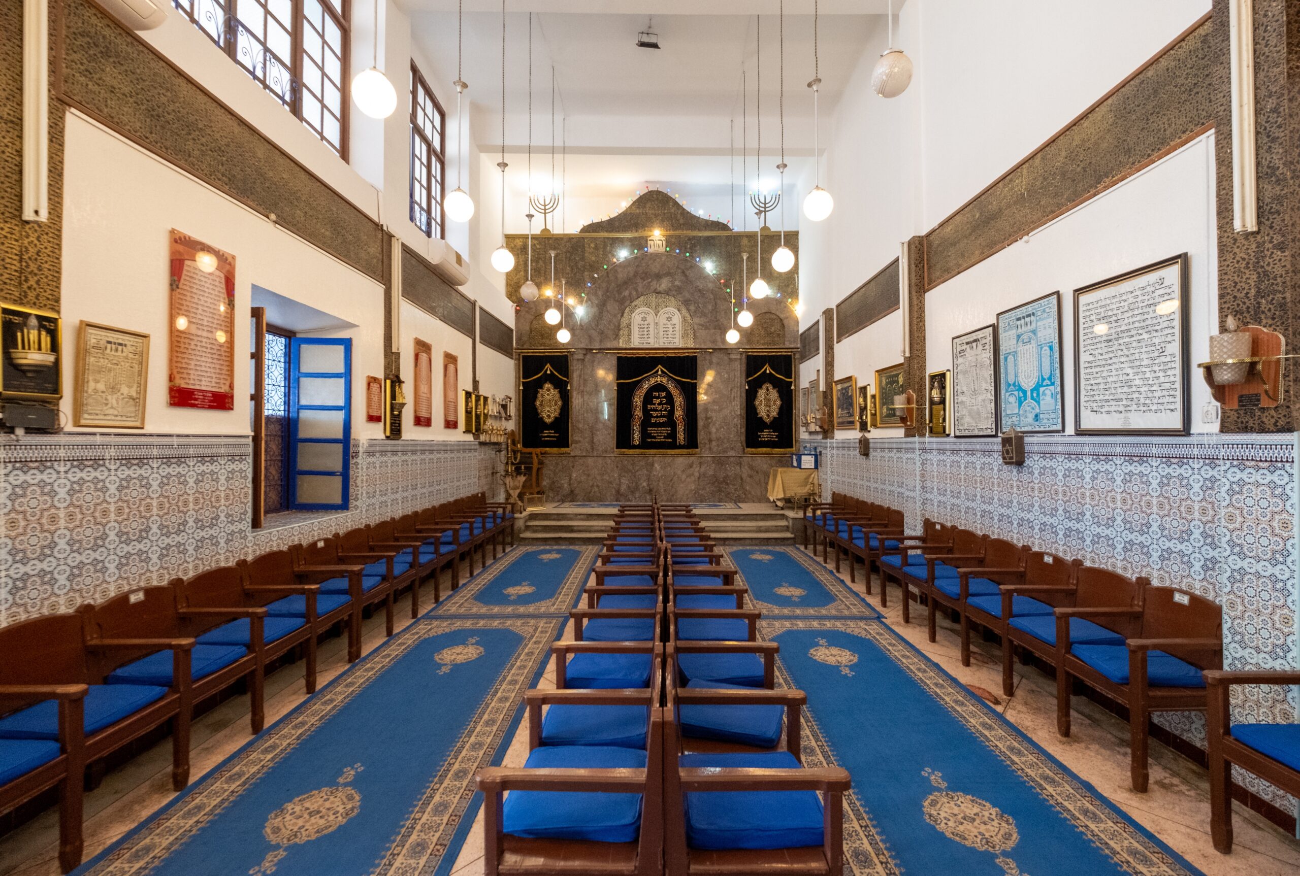 Interior of a synagogue with rows of blue-cushioned wooden benches, a long blue carpet runner, and a decorative ark at the far end.