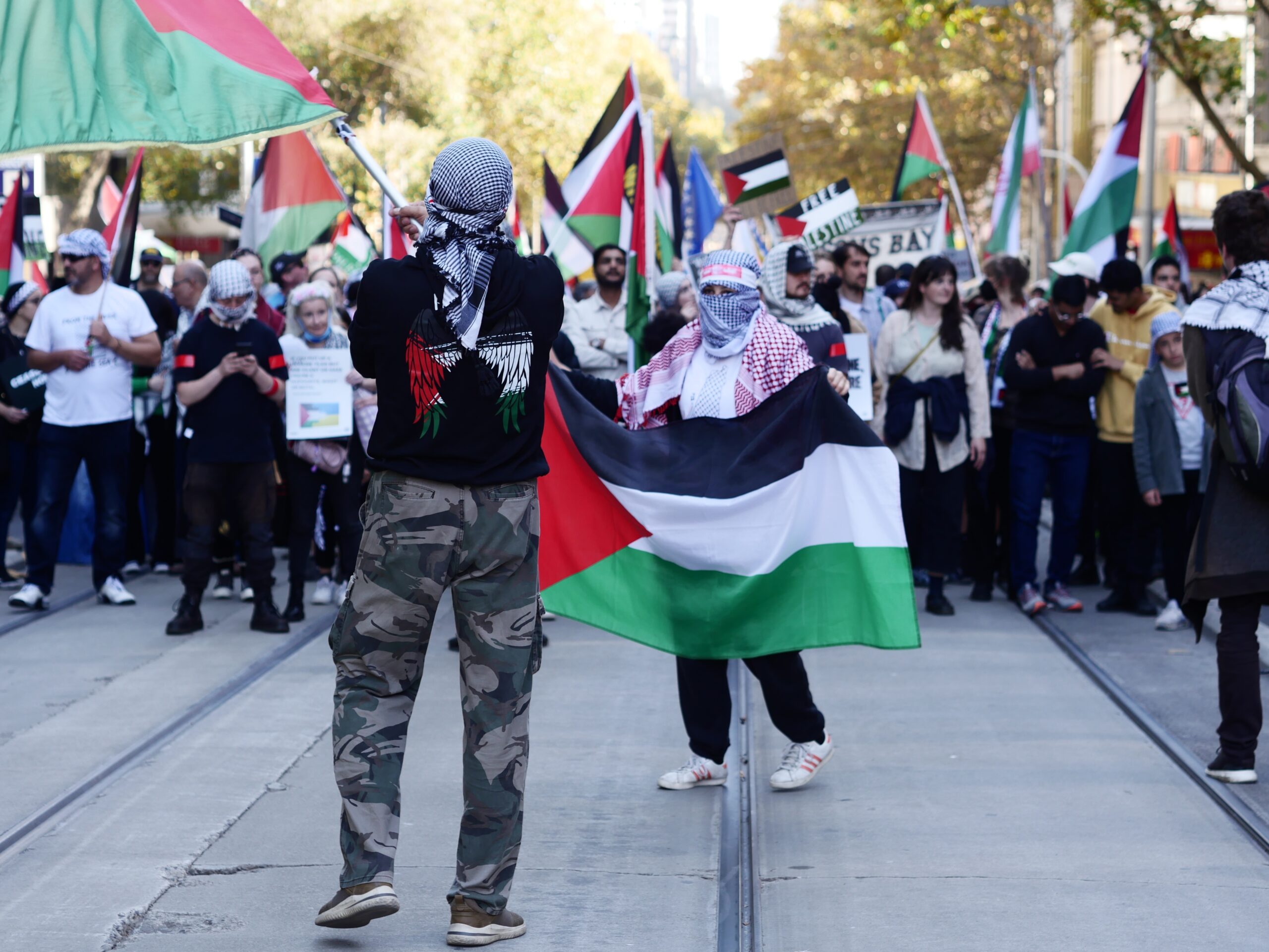 Crowd of protesters marching in a city street, many waving Palestinian flags; foreground shows a person with camouflage pants and a keffiyeh scarf facing away.