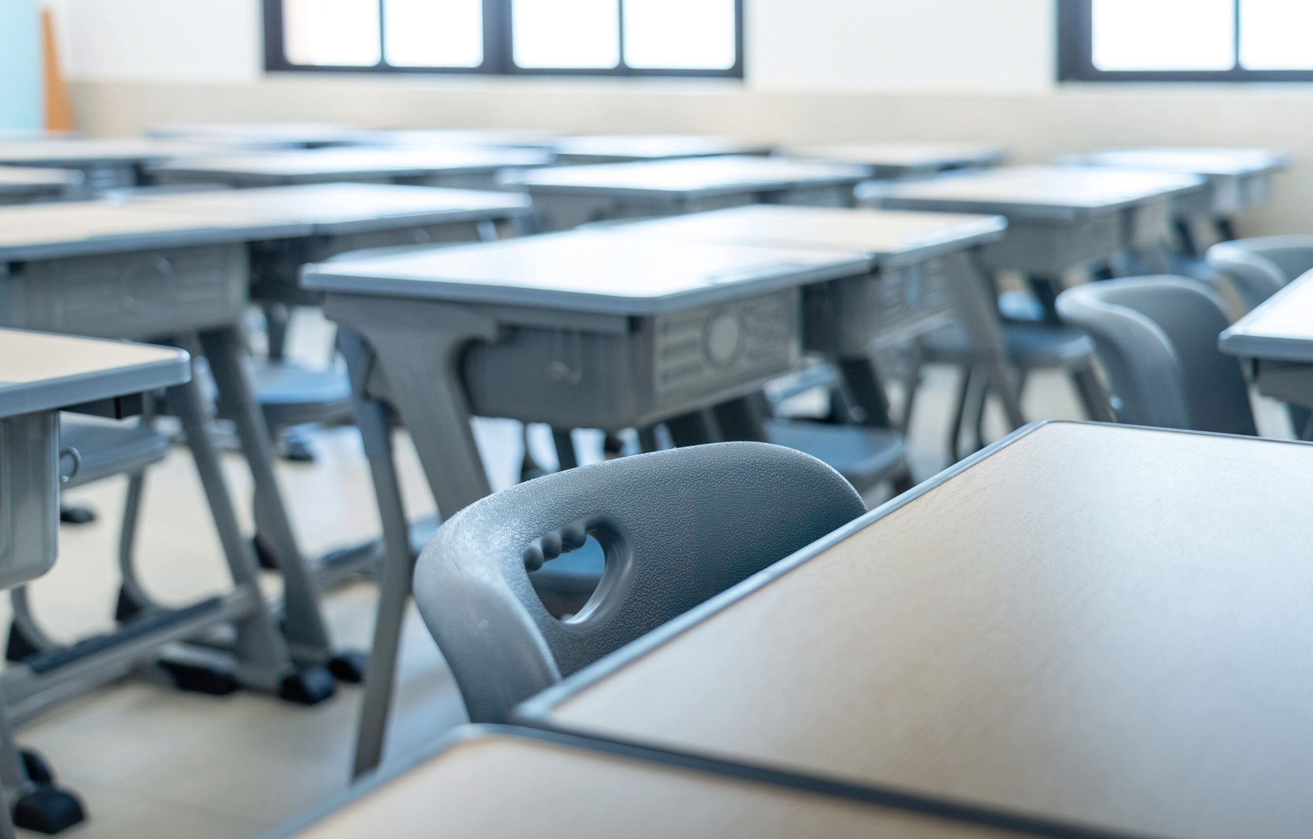 Blurred view of an empty classroom with rows of desks and chairs, seen from a close-up desk edge near the camera, windows in the background