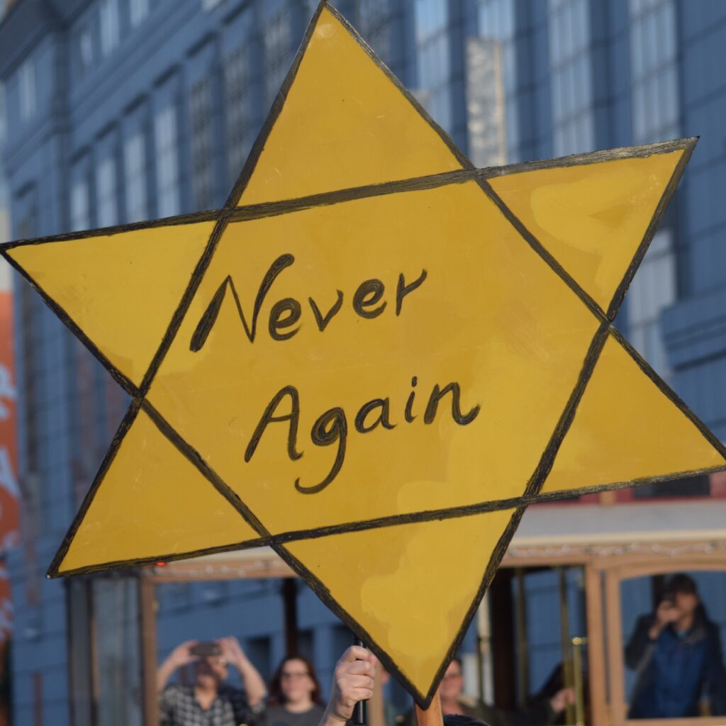 Yellow diamond protest sign held up in foreground with handwritten text, showing the word 'Never' (likely 'Never again'); blurred crowd and city buildings in background.