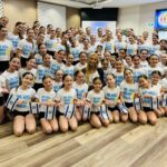 Large group of young female dancers in white 'We Will Dance Again' shirts, posing with Israeli flags in a studio.
