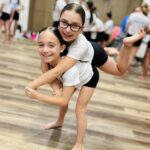 Two smiling girls in white shirts share a playful piggyback ride in a gym, with others blurred in the background.