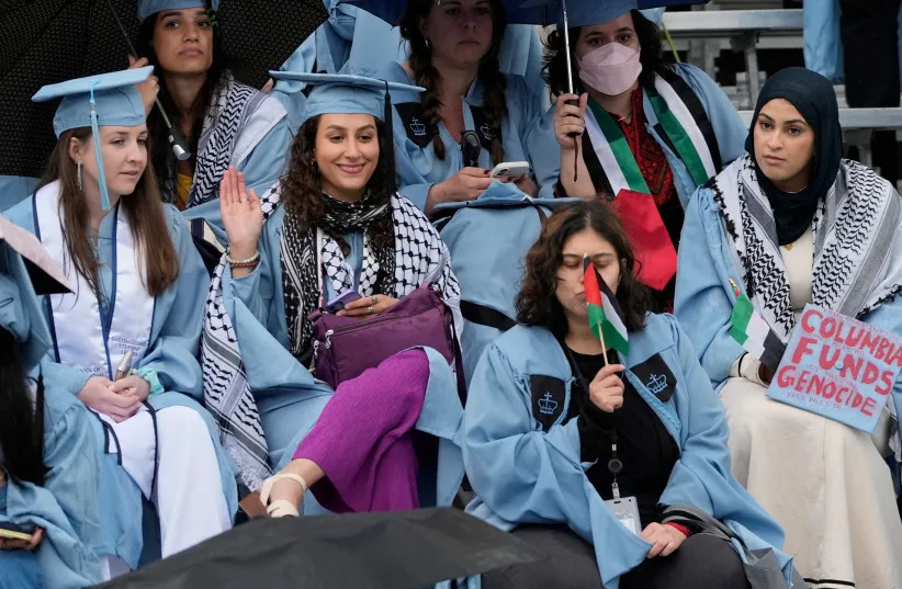 673749 - StandWithUs Diverse graduates in light-blue gowns sit in bleachers; one holds a Palestinian flag and another holds a sign that reads 'Columbia Funds Genocide'