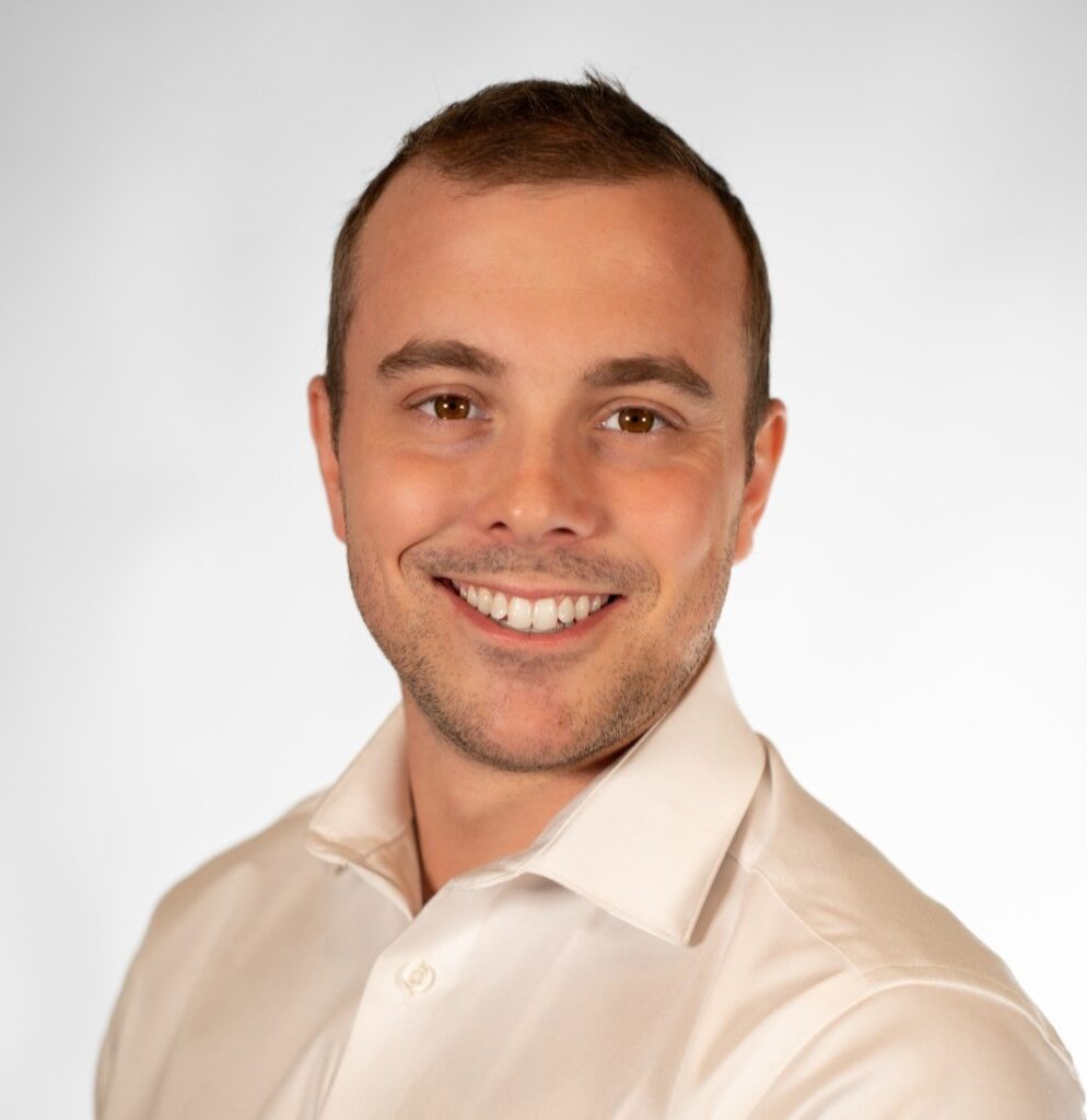 Professional headshot of a smiling man in a light beige dress shirt against a neutral gray background, facing slightly to the right.