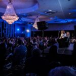 Formal gala or conference in a ballroom: audience seated at tables while two presenters speak on a raised stage under large crystal chandeliers bathed in blue light