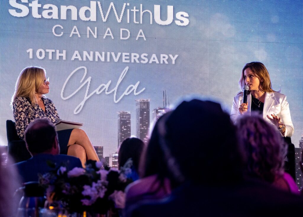 Two women seated on a stage in a panel discussion at StandWithUs Canada 10th Anniversary, backdrop shows a city skyline.