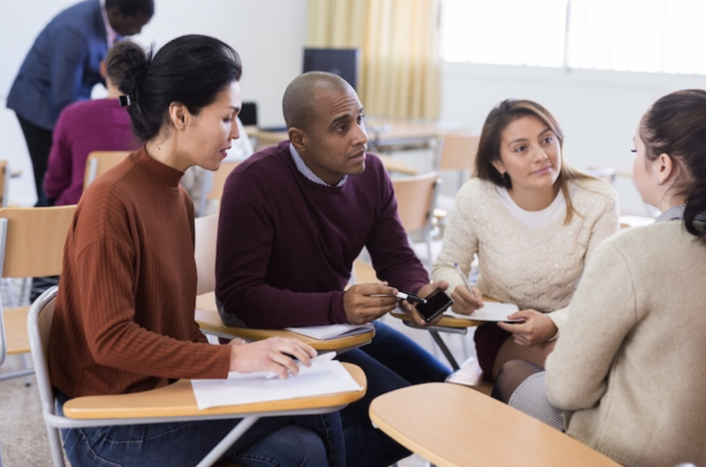 Four students in a classroom collaborate around desks, one man showing something on a phone to three others who are taking notes.