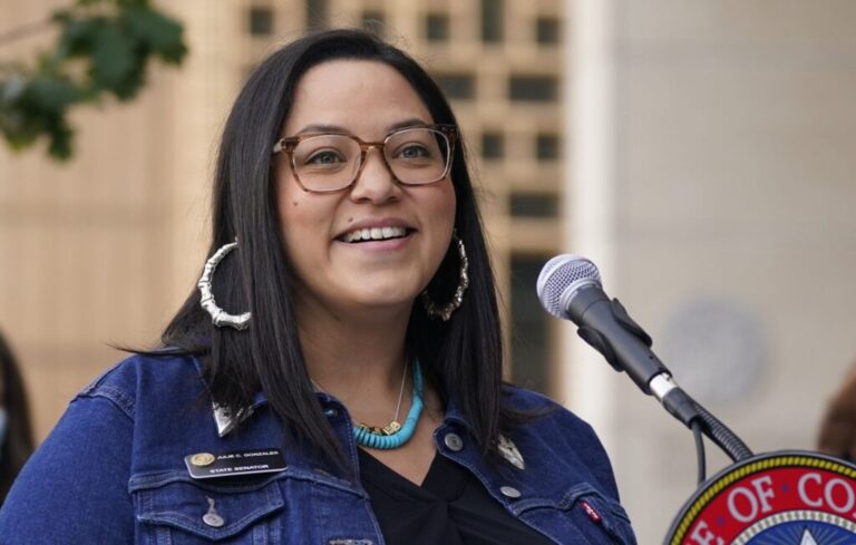 Smiling woman with glasses speaks into a microphone at a podium outdoors, wearing a denim jacket and turquoise necklace.