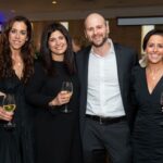 Four adults posing together at a formal event, two women on the left in black dresses, a man in a suit, and a woman on the right in black, all holding wine glasses and smiling.