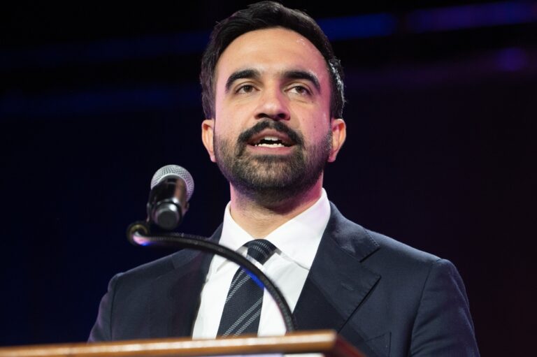 Man in a dark suit and tie speaks at a podium with a microphone during a formal event.