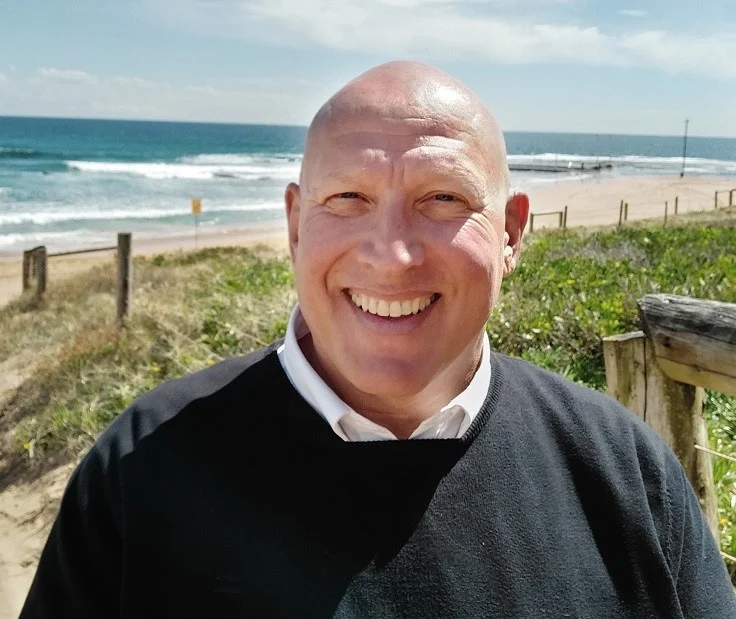Smiling bald man in a white shirt and black sweater at a sunny beach, with the ocean and a fence behind him.