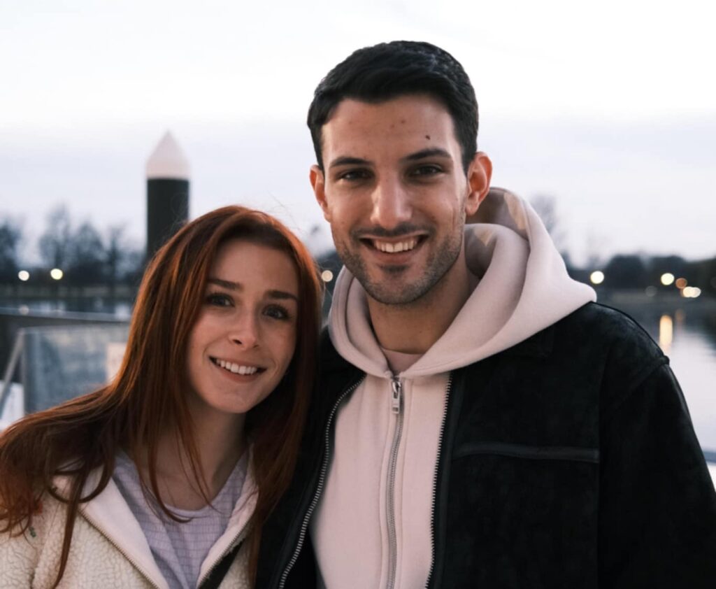 Smiling couple posing outdoors by a waterfront, with a distant tower and lights in the background.