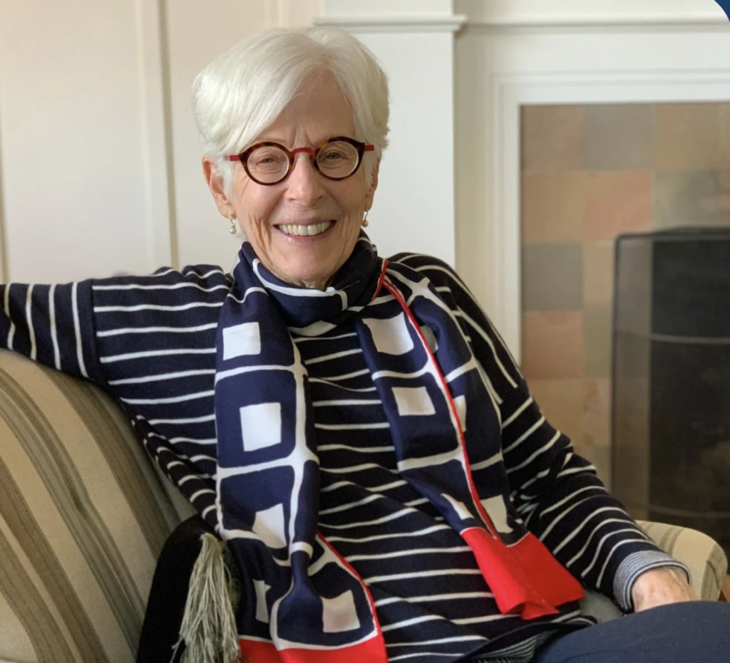 Smiling elderly woman with white hair and red glasses sits on a striped sofa, wearing a navy striped top and bold geometric scarf near a fireplace.