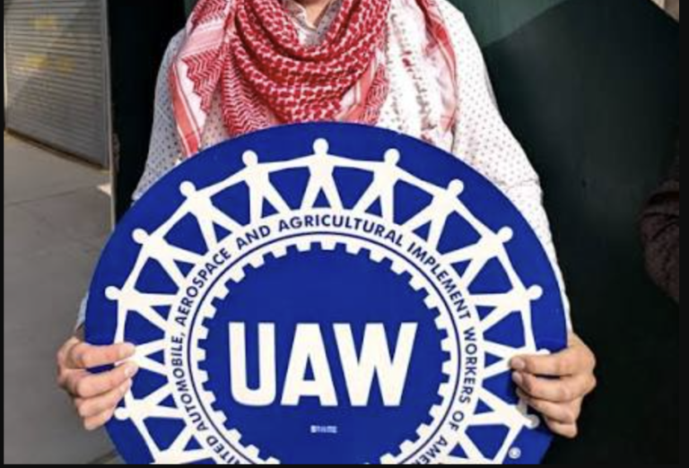 Person holding a large blue circular UAW sign featuring white worker-figures holding hands around the edge, symbolizing the autoworkers' union.