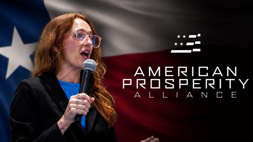 Female speaker with glasses on stage, microphone in hand, against an American flag backdrop and 'American Prosperity Alliance' logo.