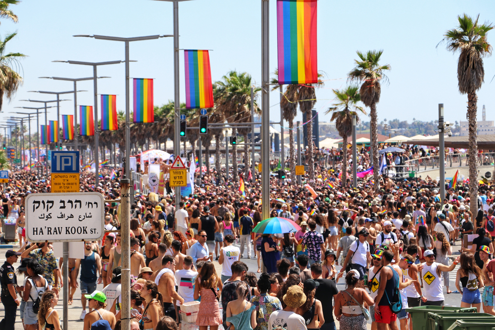 Crowded beach boardwalk during Pride parade with rainbow flags and palm trees lining the street.