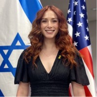 Smiling woman with long red hair in a black dress, wearing a yellow ribbon pin, standing between the Israeli and U.S. flags.