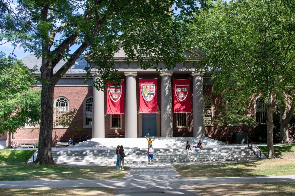 Campus scene with a brick building, white columns, and red banners hanging between the columns, framed by green trees; people sit and stand on the steps in front.