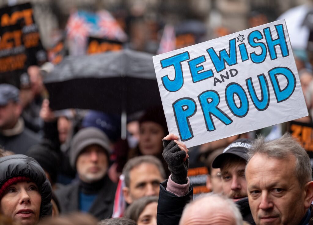 Protest crowd holds a white sign with blue letters reading 'Jewish and Proud' during a rally.