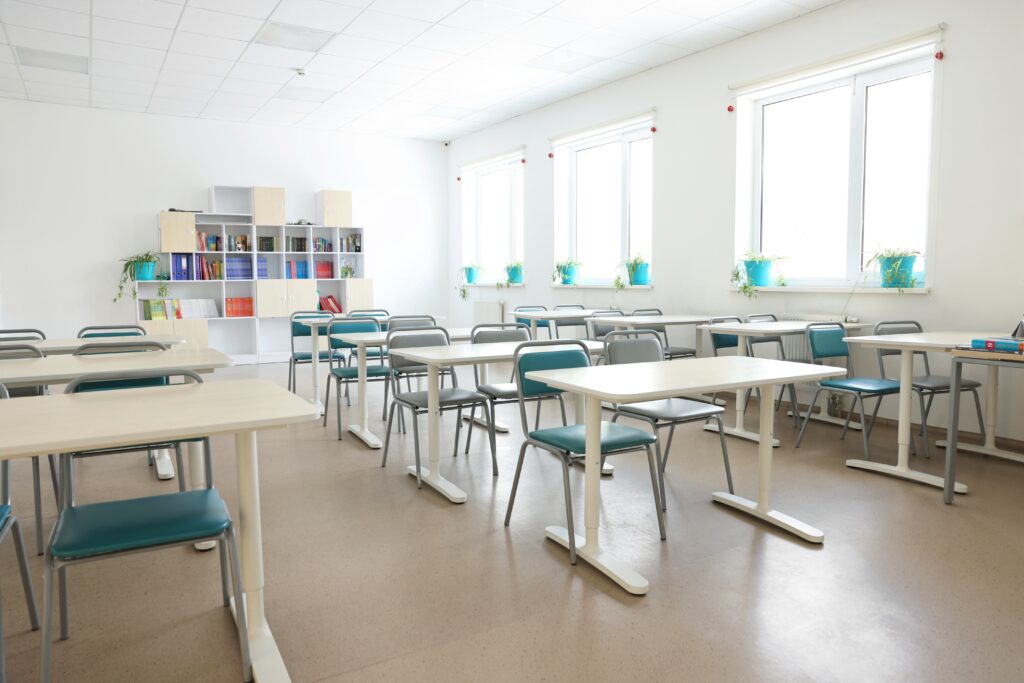 Bright classroom with white walls, rows of desks and teal chairs, and potted plants on windowsills.Natural light fills the room from large windows on one side.