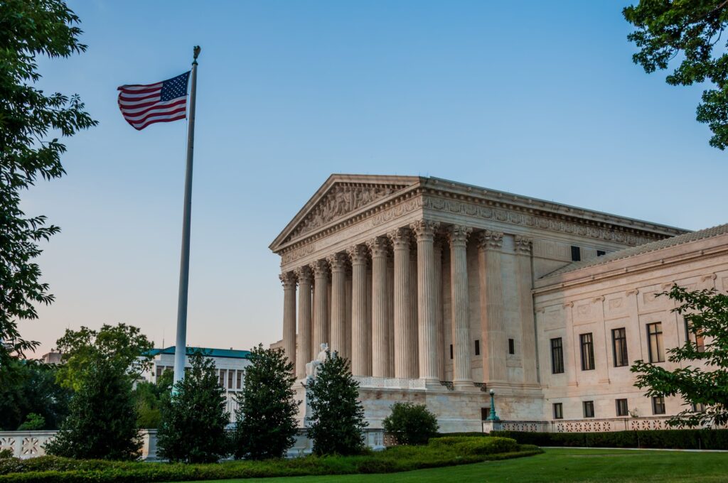American flag waving beside a neoclassical government building with tall columns against a blue sky