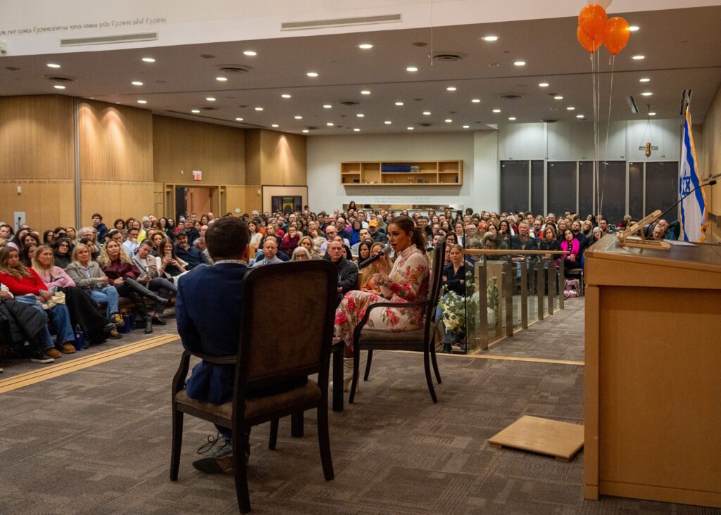 Speaker addressing a large audience in a hall with an Israeli flag and orange balloons nearby.