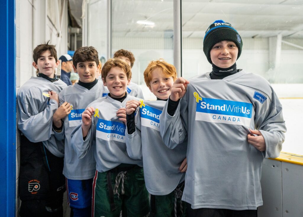 Group of smiling junior hockey players in gray jerseys standing in a rink corridor.