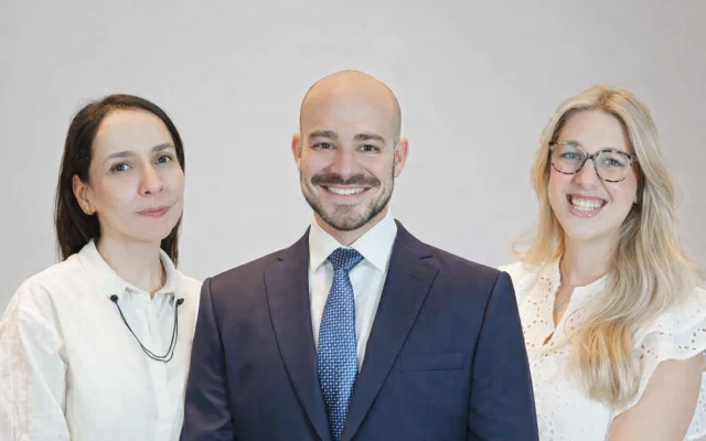 Three professionals posing together for a group photo: a man in a navy suit and blue tie flanked by two women in light-colored blouses, all smiling.