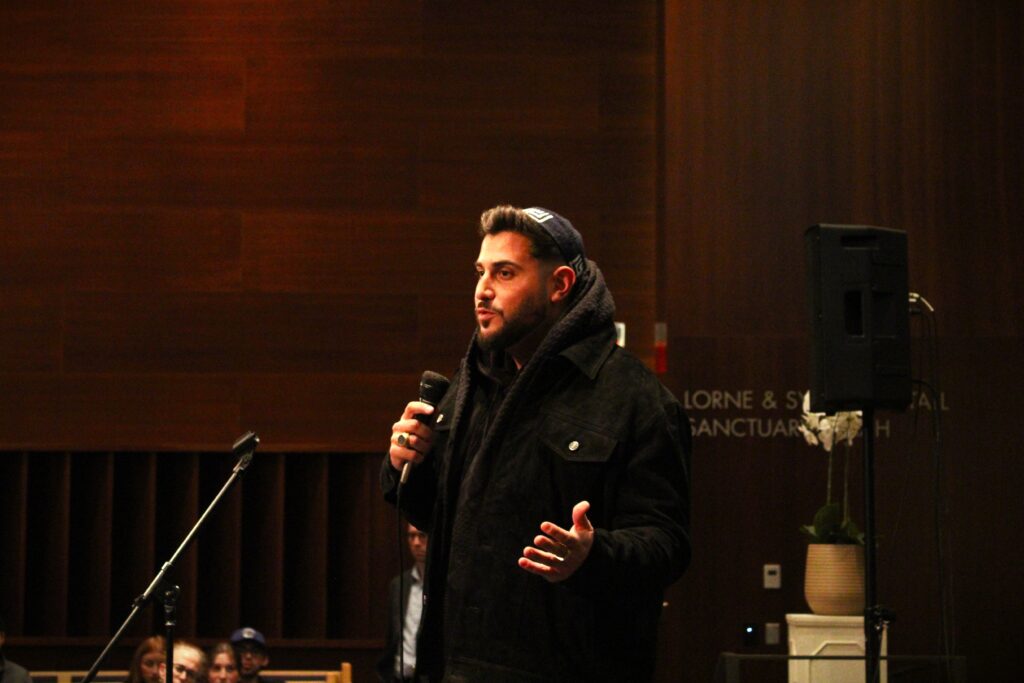 Man in a dark coat and beanie speaks into a microphone on a wood-paneled stage, with an audience in front.