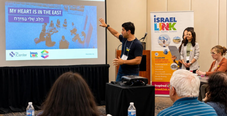 Male presenter gesturing toward a blue slide that says 'MY HEART IS IN THE EAST' during a conference; audience in foreground.