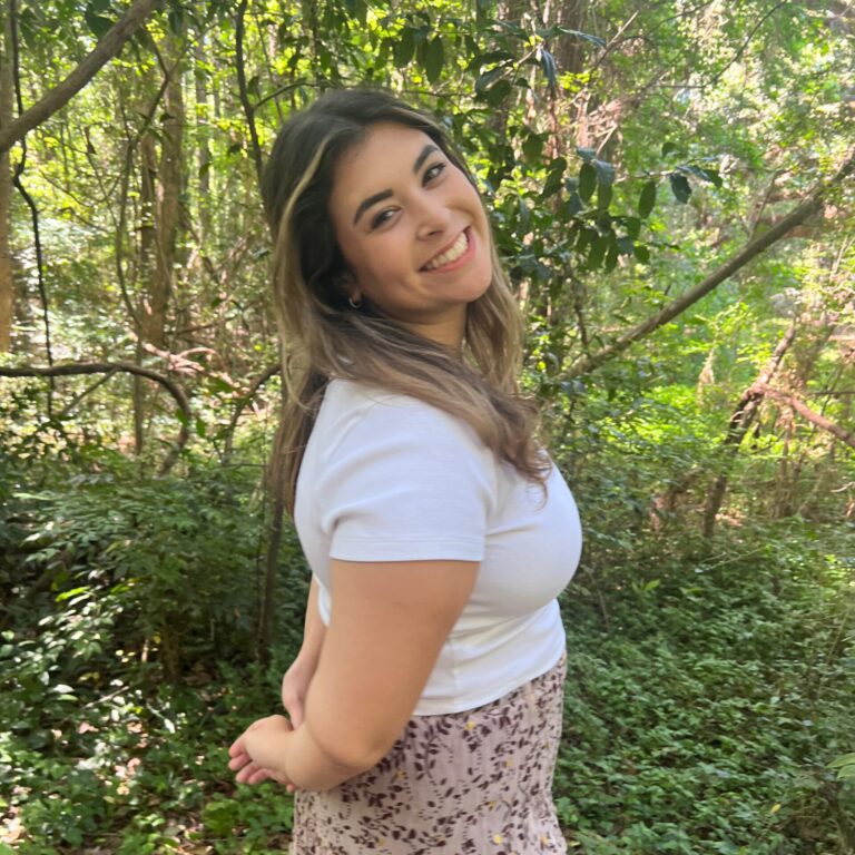 Woman smiling in a white shirt standing in a leafy forest, side view with greenery all around.