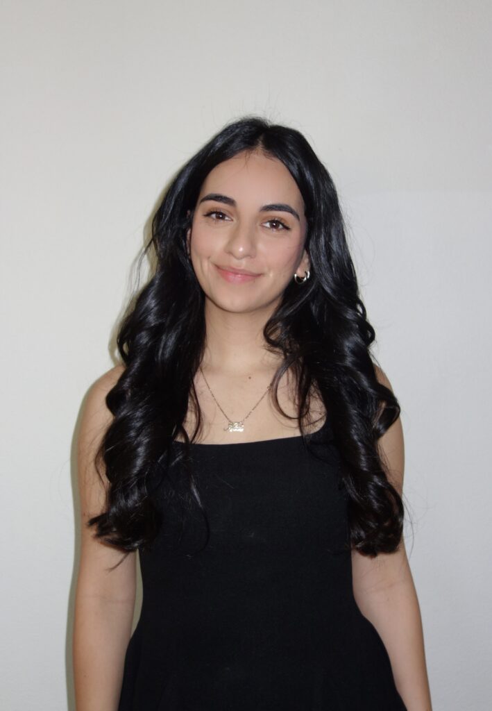 Young woman with long dark hair, smiling, wearing a black sleeveless top, standing against a light wall.