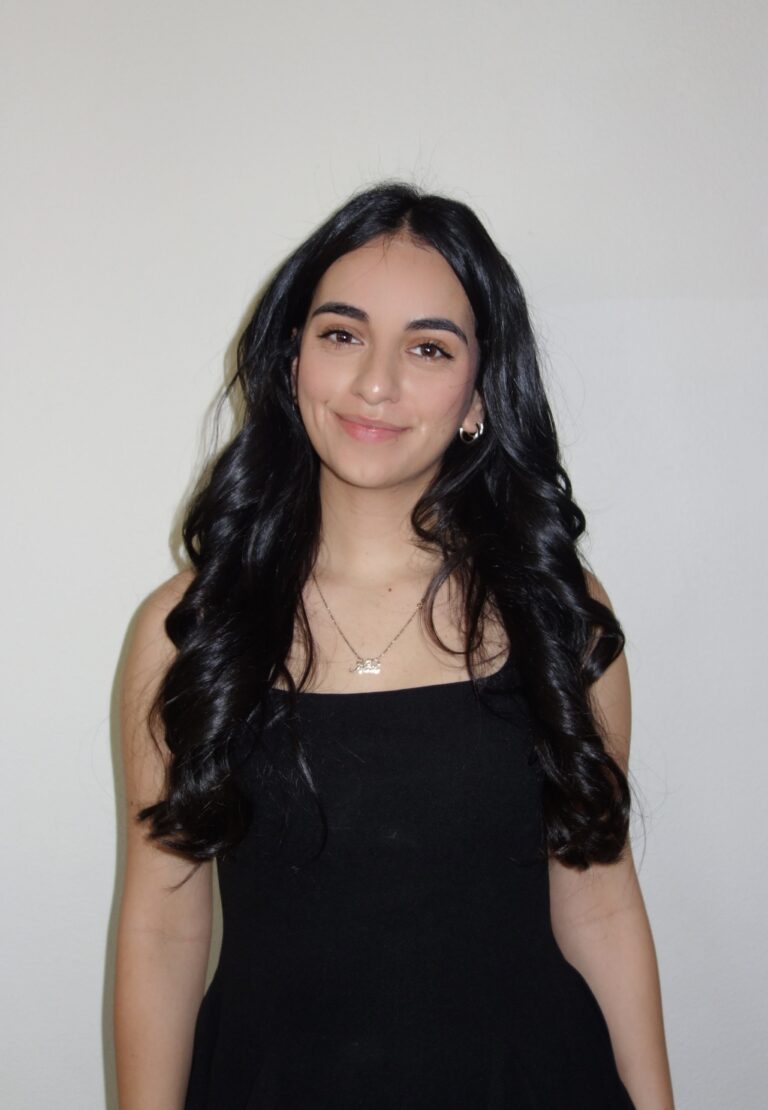 Young woman with long dark hair, smiling, wearing a black sleeveless top, standing against a light wall.