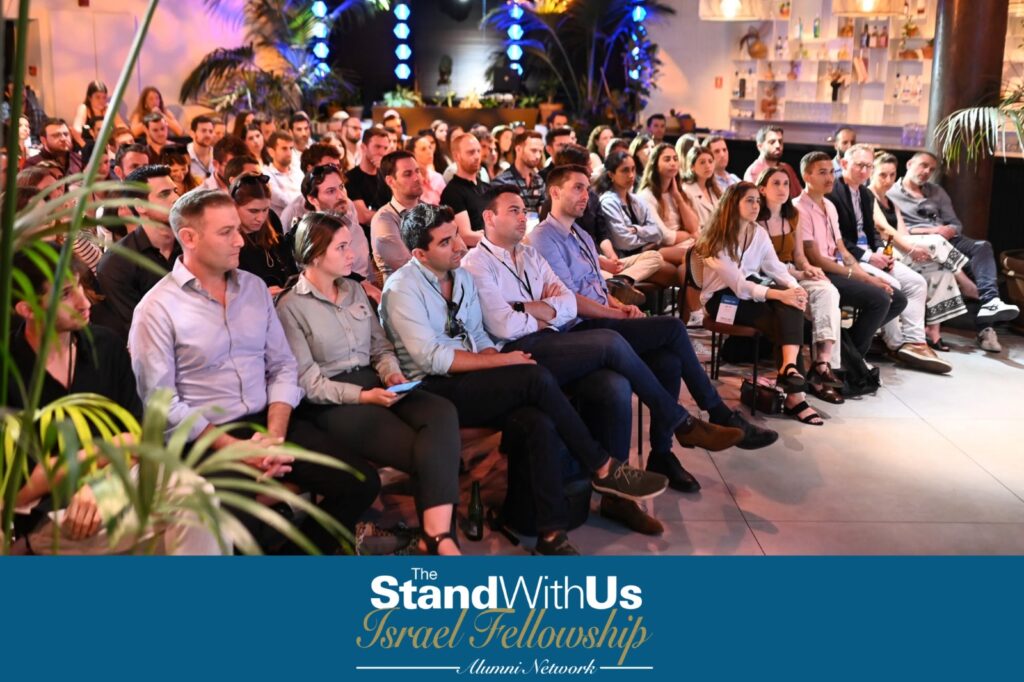 Audience seated in a conference hall listening to a speaker, with tropical plants in the background and a blue banner reading 'The StandWithUs Israel Fellowship Alumni Network'.
