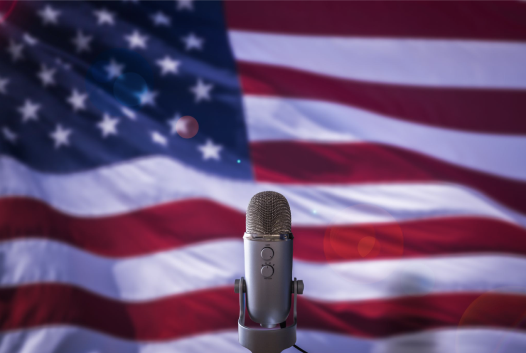 Silver microphone on a stand with a blurred American flag in the background, suggesting a patriotic podcast or speech.