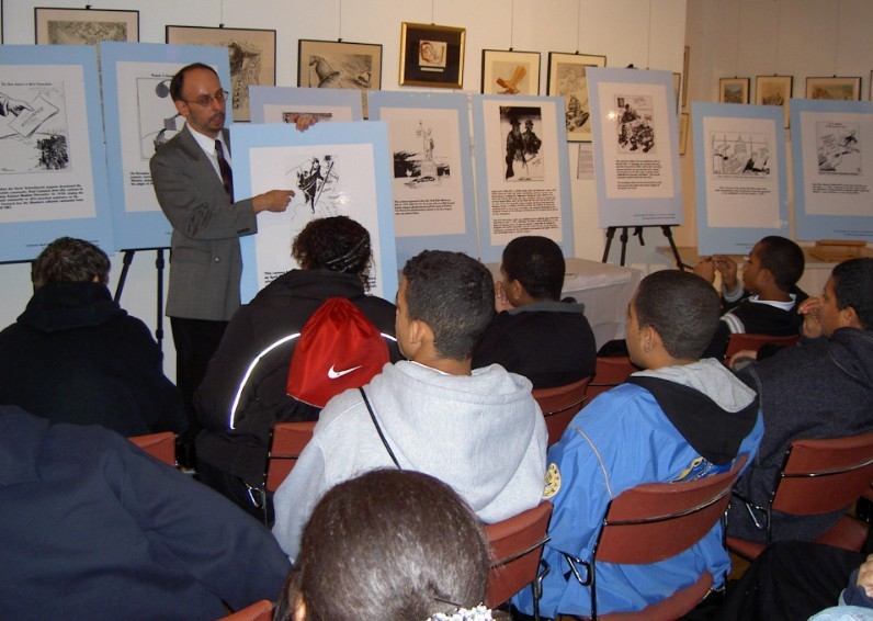 Instructor in a suit explains illustrated panels mounted on blue boards to a classroom of students in a gallery setting.